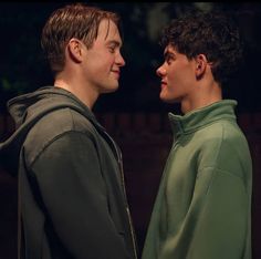 two young men standing next to each other in front of a dark background, one wearing a green hoodie