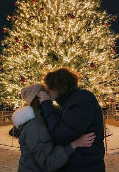 a man and woman kissing in front of a christmas tree with lights all around it