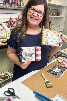 a woman holding up two small notebooks in front of her face and surrounded by craft supplies