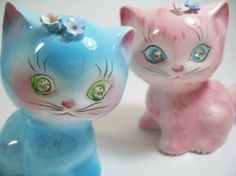 two ceramic cats sitting next to each other on a white surface with blue and pink colors