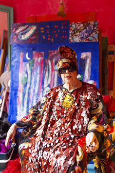 an old woman sitting in a chair with many things on the table behind her and she is wearing a colorful dress