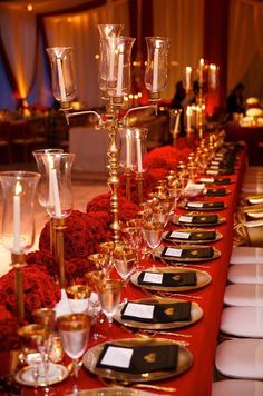 a long table is set up with gold and red plates, silverware, and candles