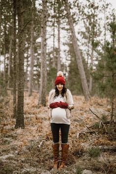 a pregnant woman standing in the woods wearing a red hat and scarf with her hands on her hips