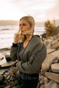 a woman standing by the water with her hand on her chin and looking off into the distance