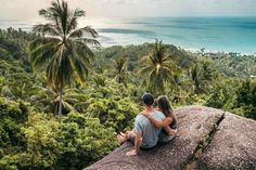 a man and woman sitting on top of a rock looking out at the ocean with palm trees