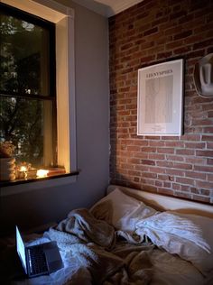 an unmade bed in front of a brick wall with a laptop computer on it