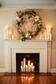 candles are lit in front of a fireplace with a wreath above it and flowers on the mantle