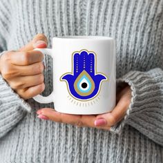 a woman holding a white coffee mug with a hamsa hand design on the front