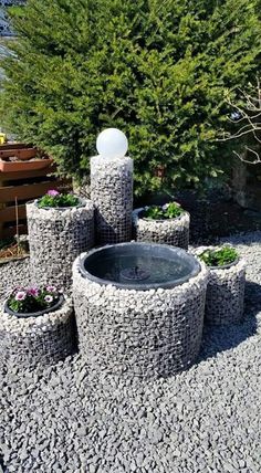 an outdoor fountain made out of rocks in the middle of a gravel area next to trees