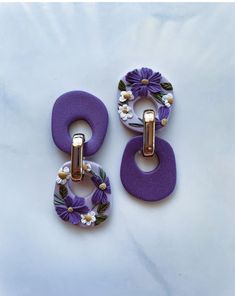 two purple and white flowered earrings on a white surface with gold hardware, one is hanging from the other side