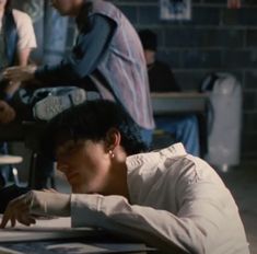 a woman sitting at a table writing on a piece of paper with other people in the background