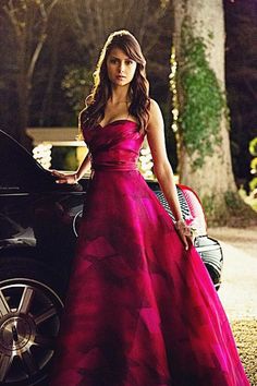 a woman in a long red dress standing next to a car and looking at the camera