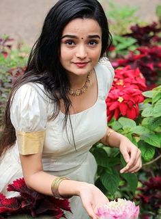 a woman is holding a pink flower in her hand and posing for the camera with red flowers behind her