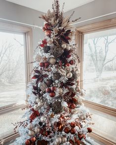 a white christmas tree with red and silver ornaments in front of two large windows on a snowy day