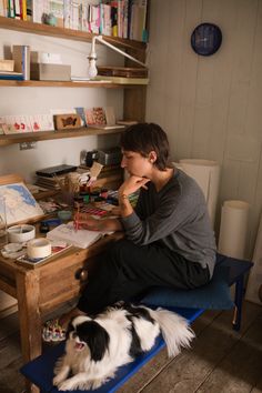 a person sitting at a desk with a dog on the floor next to it and bookshelves in the background