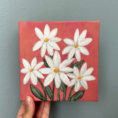 a hand is holding up a painting with white daisies on red and pink background