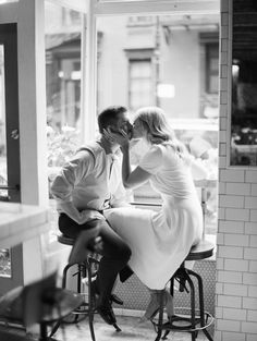 a man and woman sitting on stools in front of a store window kissing each other