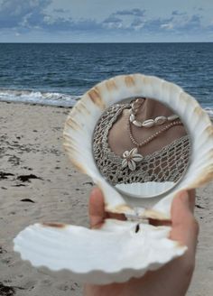 a person holding up a shell with jewelry on it at the beach in front of the ocean