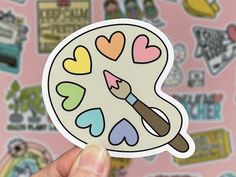 a hand holding up a sticker with hearts and a paintbrush in the center