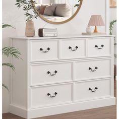 a white dresser with drawers and a round mirror on the wall above it, next to a plant