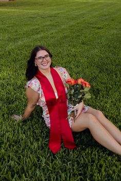 a woman sitting in the grass wearing a red graduation gown and holding a bouquet of flowers