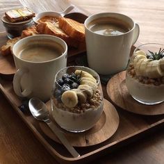 two cups of coffee with banana slices and granola in them on a wooden tray