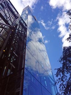 the side of a tall glass building with trees in the foreground and clouds in the background