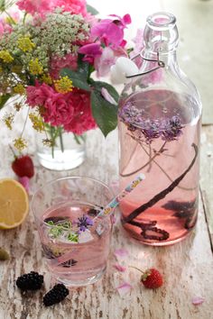 a bottle and glass filled with liquid sitting on a wooden table next to flowers, lemons and berries