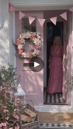 a woman standing in front of a pink door with a wreath on it and flowers