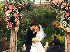 a bride and groom kissing in front of an arch with flowers on the wall behind them