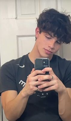 a young man looking at his cell phone in front of the mirror while wearing a black t - shirt