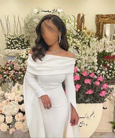 a woman standing in front of flowers wearing an off the shoulder white dress with long sleeves