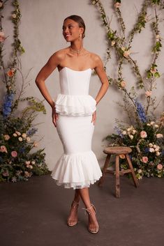 a woman wearing a white dress with ruffles on the skirt and heels standing in front of flowers