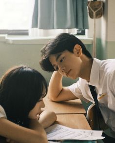 two people sitting at a table with papers and pens in front of them, one person leaning over the desk