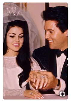 a bride and groom sitting at a table with their wedding rings on their fingers, looking at each other