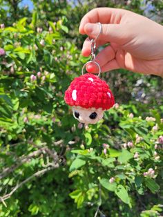 a hand holding a tiny red mushroom keychain in front of some green bushes