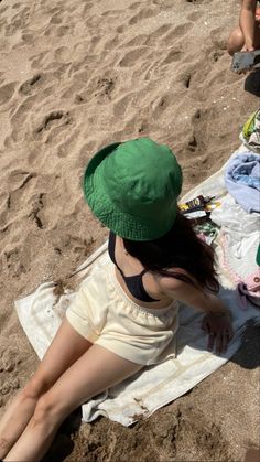 a woman in a green hat sitting on a towel at the beach with her legs crossed