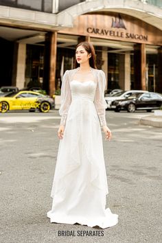 a woman standing in front of a building wearing a white dress with long sleeves and a high low neckline