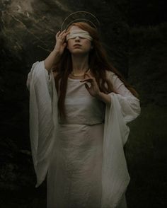 a woman with blindfolded eyes standing in front of a rock formation wearing a white dress