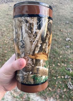 a person holding up a cup that is camouflaged with trees and grass in the background