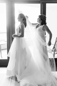 two women standing next to each other in front of a window wearing wedding gowns