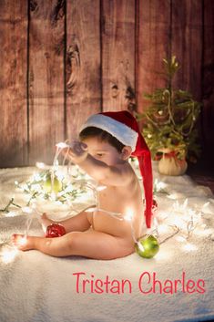 a baby wearing a santa hat sitting on top of a white blanket next to christmas lights