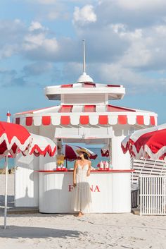 a woman standing in front of a red and white umbrella covered kiosk on the beach