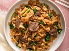 a white bowl filled with pasta and meat on top of a pink table cloth next to chopsticks