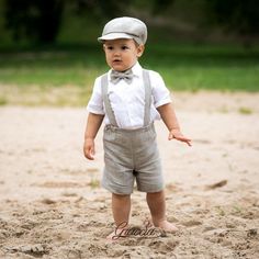 a little boy that is standing in the sand with a bow tie and suspenders