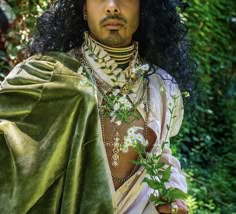 a man with long black hair is dressed in green and white clothing, holding a flower