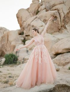 a woman in a pink dress standing on top of a rock formation with her arms outstretched