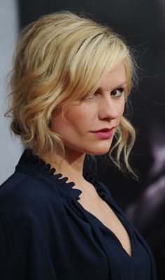 a woman with blonde hair wearing a black top and looking off to the side while standing in front of a movie poster