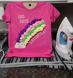 a pink shirt that says 100 days brighter next to an ironing board with a rainbow on it