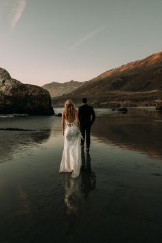 a bride and groom walking on the beach at sunset, with mountains in the background